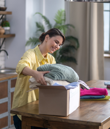 Smiling woman in a yellow shirt packing neatly folded sweaters into a cardboard donation box on a wooden table at home.
