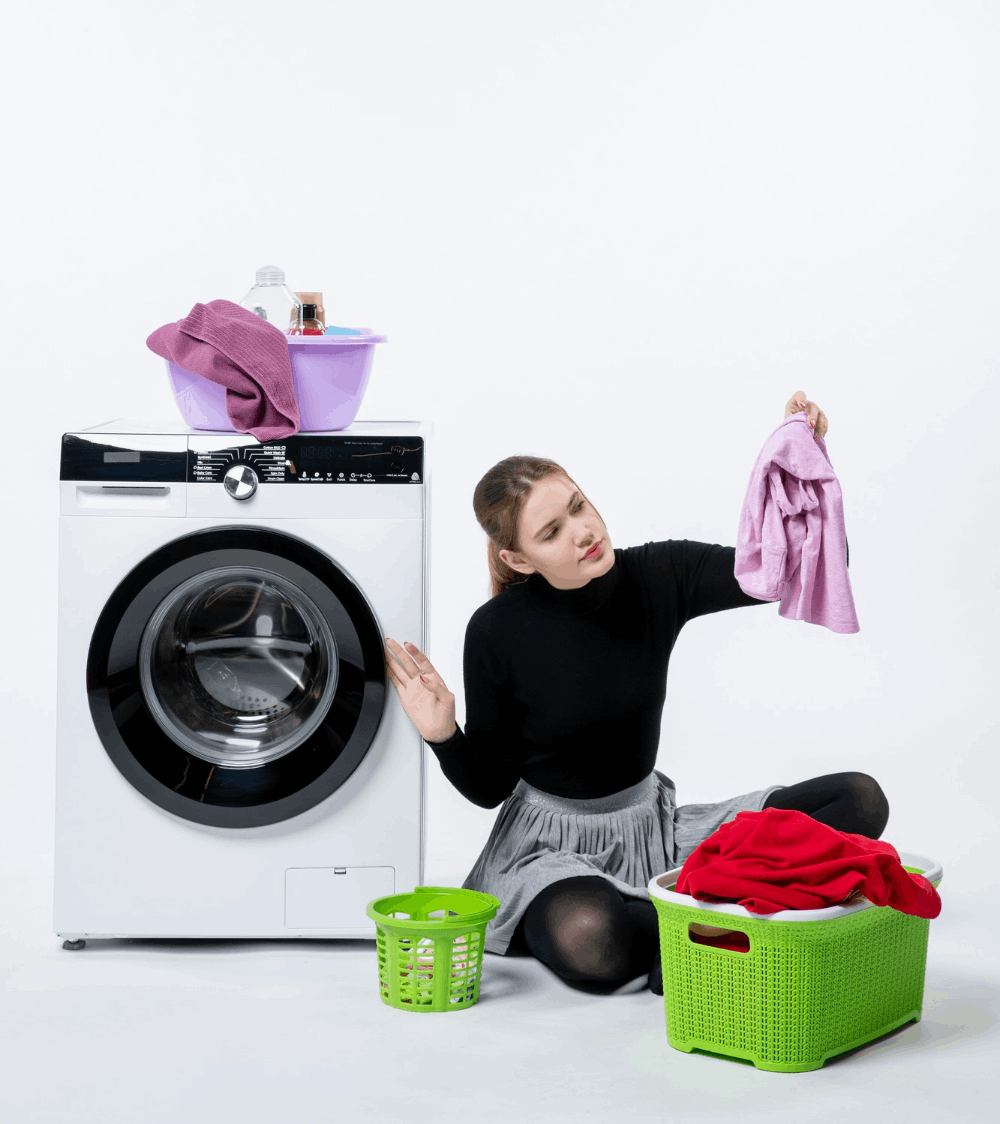 Young woman sitting on the floor next to a washing machine, examining a pink shirt while sorting laundry